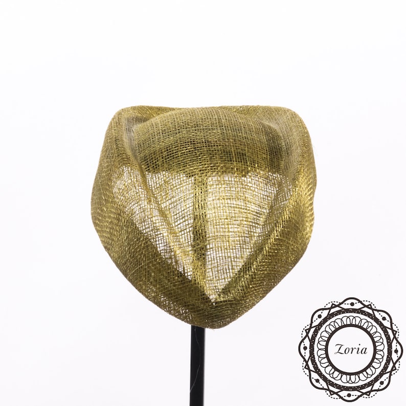 Op de afbeelding: Een goudkleurige fascinator-hoed met een gestructureerde, open geweven gaastextuur en een gebogen, sculpturaal silhouet. Het stuk is gemonteerd op een dunne zwarte standaard en heeft een rond logo in de rechterbenedenhoek met de tekst Zoria.