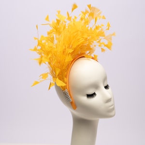May include: A yellow feather headband with a satin band and rhinestone embellishments. The headband is worn on a white mannequin head.