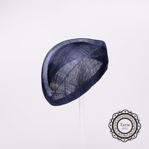 Op de afbeelding: Een marineblauwe fascinator hoed met een gestructureerd, gebogen silhouet en een zichtbare geweven textuur. De hoed heeft een opvallend, omhoogstaand randontwerp dat een sculpturale vorm cre&euml;ert. Een cirkelvormig logo met de tekst Zoria is zichtbaar.