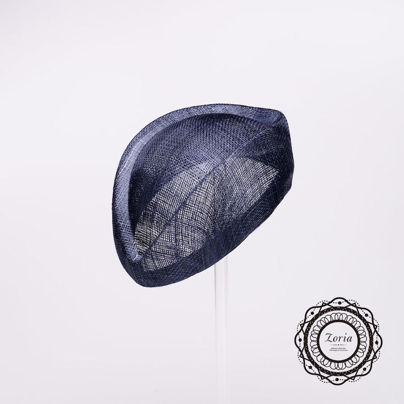 Op de afbeelding: Een marineblauwe fascinator hoed met een gestructureerd, gebogen silhouet en een zichtbare geweven textuur. De hoed heeft een opvallend, omhoogstaand randontwerp dat een sculpturale vorm cre&euml;ert. Een cirkelvormig logo met de tekst Zoria is zichtbaar.