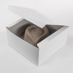 May include: A tan knit hat rests inside a white, open box. The hat has a textured, woven appearance. The box is rectangular with a partially open lid, revealing the hat inside. The background is plain white.