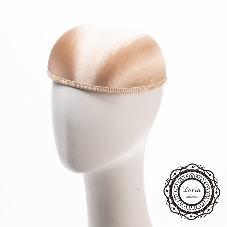 May include: A dome-shaped pillbox hat in a smooth, champagne-coloured satin finish. The hat features a structured, rounded crown and is finished with a textured, woven trim along the bottom edge. A circular logo with the text Zoria is visible.