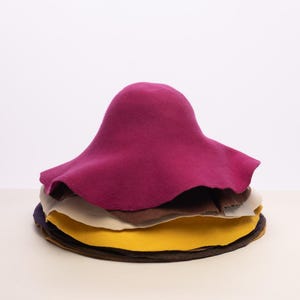 May include: A stack of felt hats in various colors, including a vibrant magenta, brown, yellow, and navy. The top hat is magenta with a wide brim. The hats are stacked on top of each other.