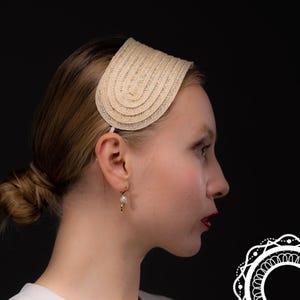 May include: A woman with blonde hair is wearing a beige headband with a woven design. She is wearing a pearl earring.