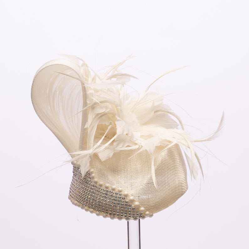 Op de afbeelding: Een ivoorkleurige fascinator met een gestructureerde basis versierd met een band van sprankelende heldere strass-steentjes en een rij kleine witte parels. De bovenkant is gedecoreerd met een grote, golvende lus van stijve stof en zachte veren.