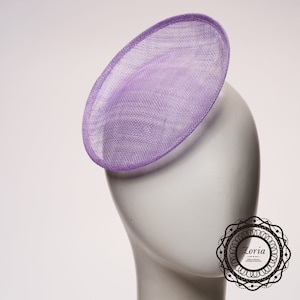 May include: A lavender fascinator hat with a circular, slightly tilted design and a woven, mesh-like texture. The hat is displayed on a plain white mannequin head. A circular logo with the text Zoria Millinery Materials Trimming & Accessories is visible in the bottom right corner.
