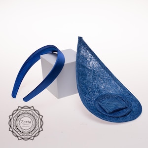 May include: A royal blue satin headband paired with a matching teardrop-shaped fascinator base made of a textured, woven material. The fascinator features a circular attachment point. A logo with the text Zoria is visible in the bottom corner.