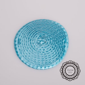 May include: A circular, light blue woven ribbon accessory featuring a coiled design with a smooth, shiny texture. In the bottom right corner, a black circular logo contains the text Zoria, followed by smaller text reading Millinery Materials, Trimmings & Accessories.