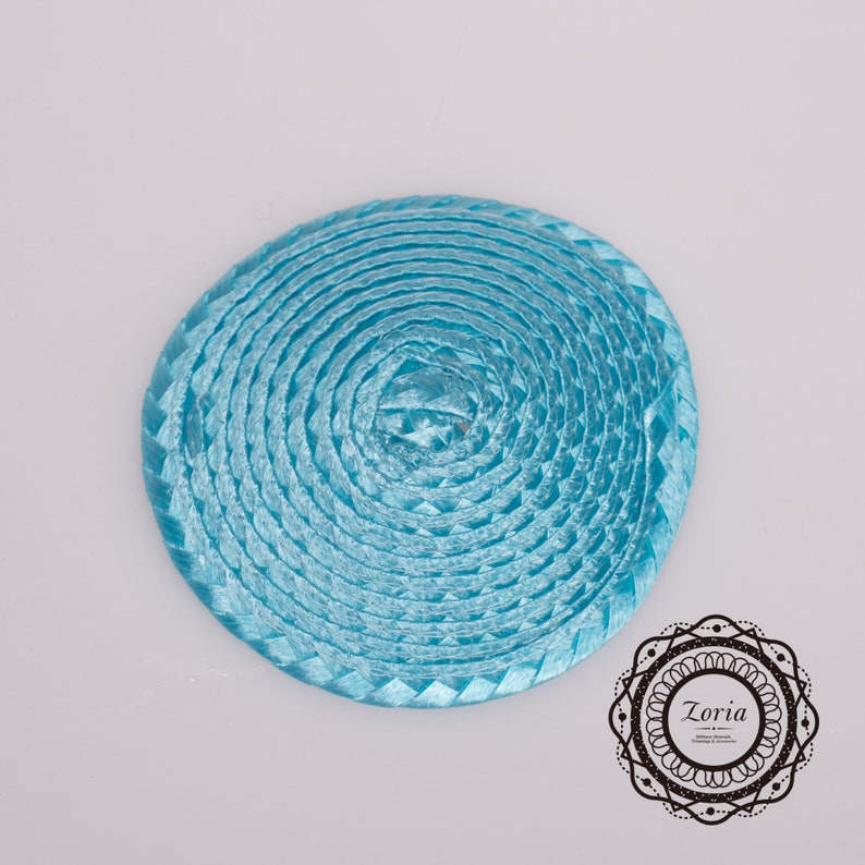 May include: A circular, light blue woven ribbon accessory featuring a coiled design with a smooth, shiny texture. In the bottom right corner, a black circular logo contains the text Zoria, followed by smaller text reading Millinery Materials, Trimmings & Accessories.