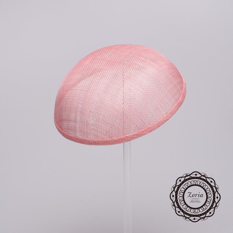 May include: A dome-shaped pink fascinator hat crafted from a woven, semi-sheer material with a structured brim edge. The hat features a smooth, rounded silhouette and is displayed on a clear stand. A circular logo with the text Zoria is visible.