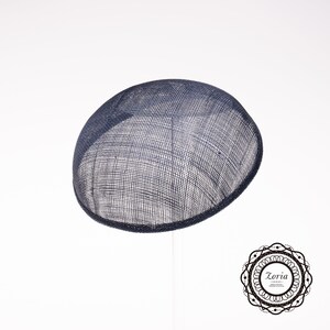 May include: A dark navy blue circular fascinator hat crafted from a stiffened, woven mesh material with a visible grid texture. The hat features a smooth, rounded dome shape and is positioned on a thin, vertical support stand. A logo with the text Zoria is visible.