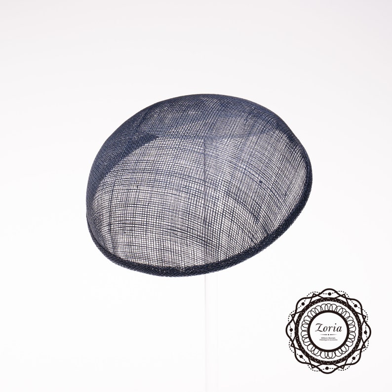 May include: A dark navy blue circular fascinator hat crafted from a stiffened, woven mesh material with a visible grid texture. The hat features a smooth, rounded dome shape and is positioned on a thin, vertical support stand. A logo with the text Zoria is visible.