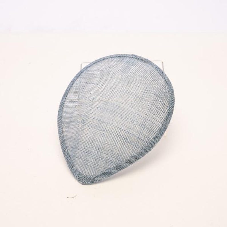 May include: A teardrop-shaped fascinator hat crafted from a light blue, fine-mesh woven material. The accessory features a structured, reinforced edge border in a matching light blue tone, creating a clean and defined silhouette for this headpiece.