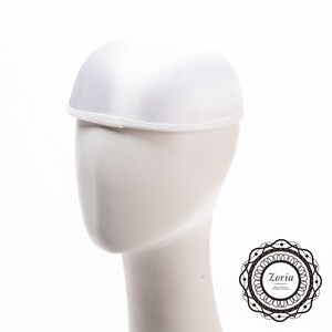 May include: A solid white dome-shaped hat with a smooth satin-like finish and a narrow, textured white trim along the bottom edge. The hat is displayed on a plain white mannequin head, featuring a clean, rounded silhouette and a simple design.