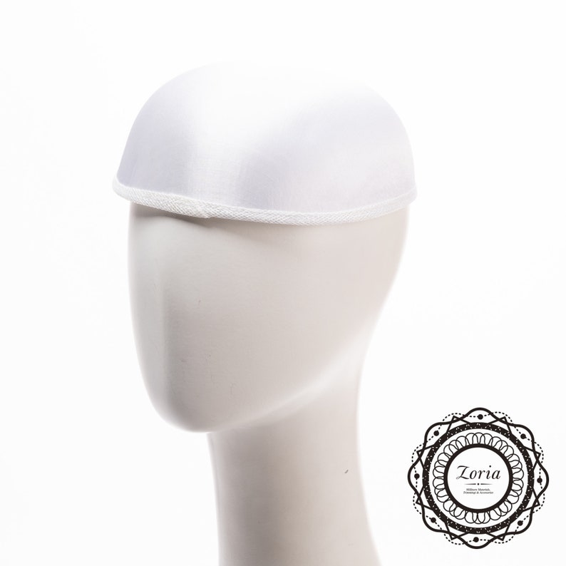 May include: A solid white dome-shaped hat with a smooth satin-like finish and a narrow, textured white trim along the bottom edge. The hat is displayed on a plain white mannequin head, featuring a clean, rounded silhouette and a simple design.