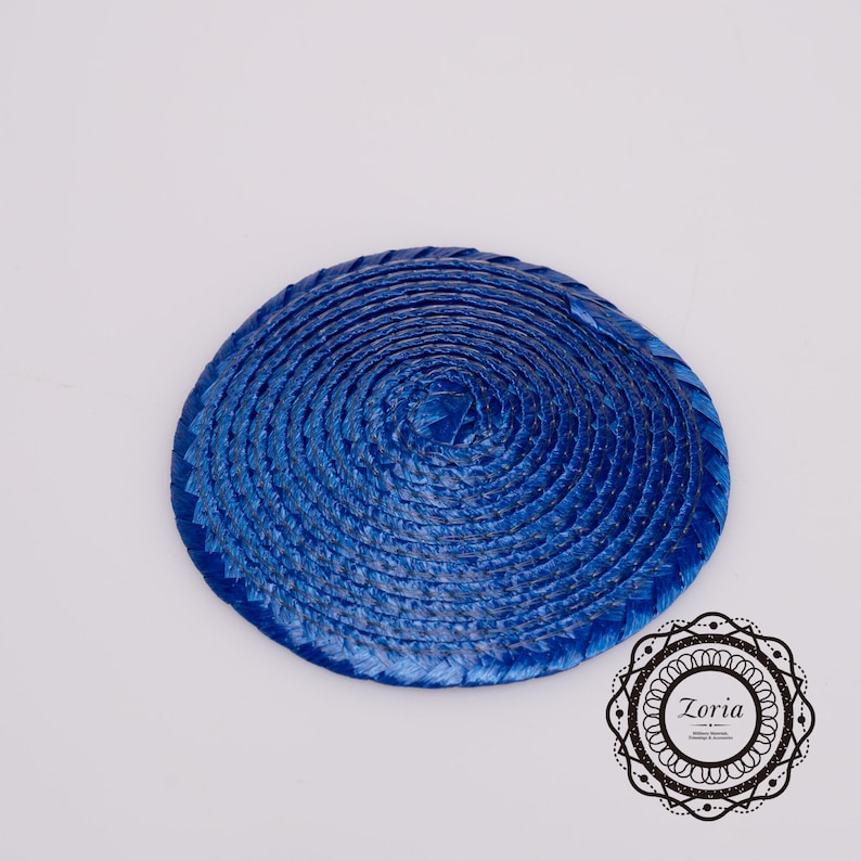 May include: A circular, royal blue fascinator base crafted from tightly coiled, woven straw-like material. The piece features a smooth, spiraling texture that radiates from the center to the outer edge. A logo with the text Zoria is visible.