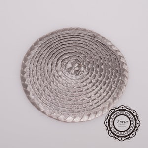 May include: A circular, silver-toned braided cord coaster or mat featuring a continuous spiral pattern. In the bottom right corner, there is a decorative black circular logo containing the text Zoria and smaller illegible script.