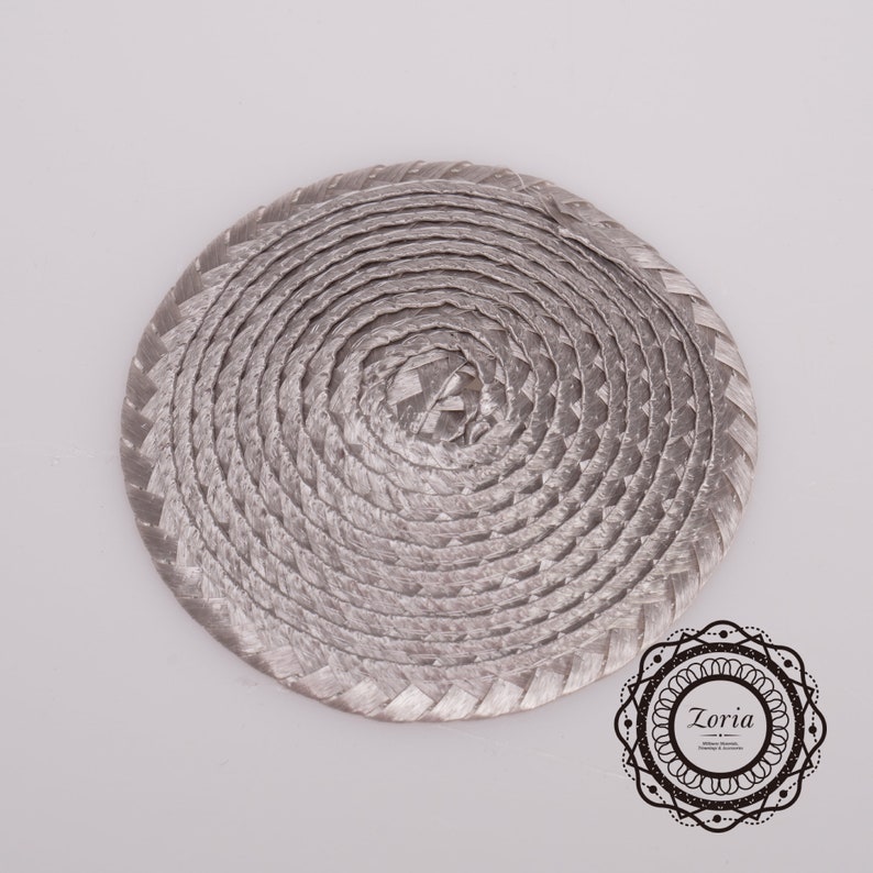 May include: A circular, silver-toned braided cord coaster or mat featuring a continuous spiral pattern. In the bottom right corner, there is a decorative black circular logo containing the text Zoria and smaller illegible script.