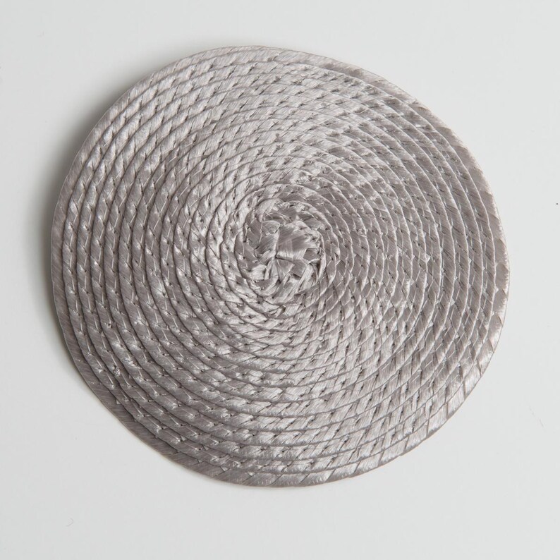 May include: A circular coaster featuring a light gray, metallic-toned rope coiled in a continuous spiral pattern. The rope is tightly wound from the center outward, creating a textured, concentric design with a subtle sheen throughout.