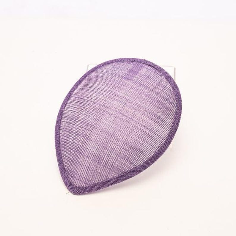 May include: A teardrop-shaped fascinator base in a vibrant purple hue, featuring a fine, woven mesh texture throughout. The piece is finished with a matching purple bound edge that defines its distinct, curved silhouette and structured form.
