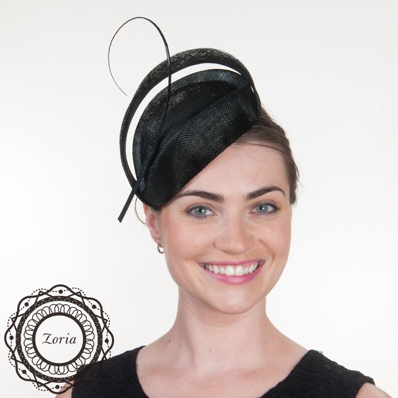 Op de afbeelding: Een zwarte fascinator met een cirkelvormige, getextureerde basis met een gebogen, verhoogde rand en een dun, zwierig veeraccent. Het ontwerp bevat een cirkelvormig logo in de linkerbenedenhoek met de tekst Zoria in een decoratieve rand.