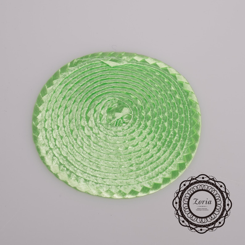 May include: A coiled length of light green woven ribbon arranged in a flat circular spiral shape. The ribbon has a shiny, textured finish with a repeating braided pattern. A circular logo with the text Zoria is visible in the bottom right.