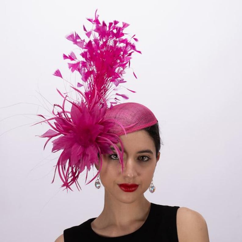 May include: A vibrant fuchsia fascinator hat featuring a structured, rounded base adorned with a large, layered floral arrangement made of matching feathers and delicate, upright plumes that extend vertically from the side of the headpiece.
