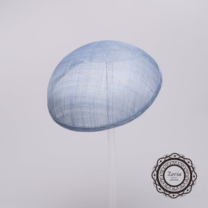 May include: A light blue fascinator hat with a woven, grid-like texture and a rounded, dome-shaped crown. The hat features a clean, structured edge and is displayed on a clear stand. A circular logo with the text Zoria is in the corner.