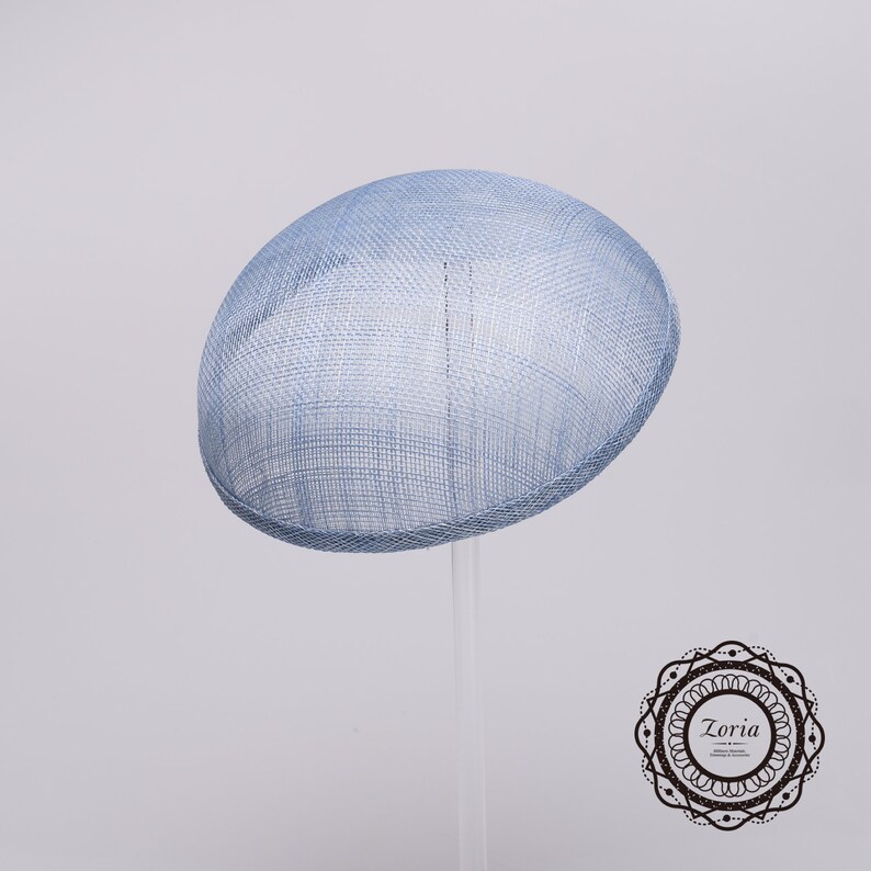 May include: A light blue fascinator hat with a woven, grid-like texture and a rounded, dome-shaped crown. The hat features a clean, structured edge and is displayed on a clear stand. A circular logo with the text Zoria is in the corner.