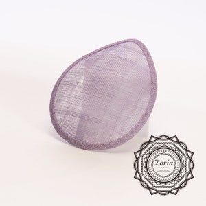 May include: A teardrop-shaped lavender fascinator base crafted from stiffened sinamay straw mesh with a matching bound edge. The accessory features a fine, woven texture and is positioned at an angle. A black circular logo with the text Zoria is visible.
