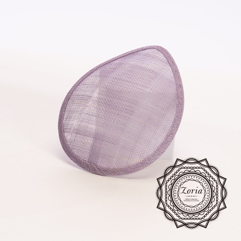 May include: A teardrop-shaped lavender fascinator base crafted from stiffened sinamay straw mesh with a matching bound edge. The accessory features a fine, woven texture and is positioned at an angle. A black circular logo with the text Zoria is visible.