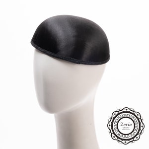 May include: A black dome-shaped pillbox hat with a smooth satin-like finish and a textured dark grey trim along the bottom edge. The hat is displayed on a plain white mannequin head, featuring a circular logo with the text Zoria in the corner.
