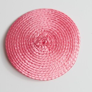 May include: A circular, flat coaster or mat constructed from a continuous coil of braided pink material. The design features a tight, spiral pattern radiating from the center outward to the edge, creating a uniform, textured surface.