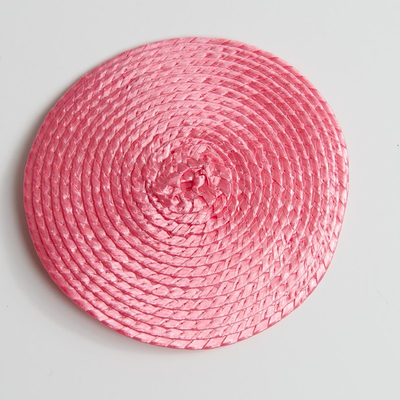 May include: A circular, flat coaster or mat constructed from a continuous coil of braided pink material. The design features a tight, spiral pattern radiating from the center outward to the edge, creating a uniform, textured surface.