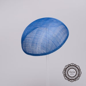 May include: A bright blue, oval-shaped fascinator hat made of a woven, mesh-like material. The hat features a structured, rounded crown and a clean, finished edge. A circular logo with the text Zoria is visible in the bottom right corner.