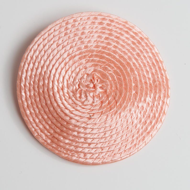 May include: A circular, light peach-colored coaster or decorative mat constructed from a continuous, tightly coiled satin cord. The surface features a concentric spiral pattern that radiates from the center to the outer edge.