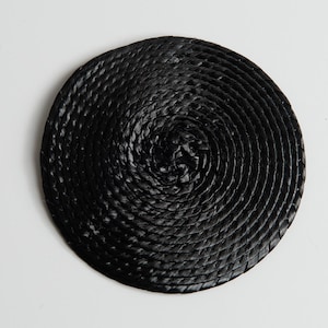 May include: A circular coaster constructed from a continuous black cord coiled in a tight spiral pattern. The surface features a textured, braided appearance created by the overlapping layers of the dark material, forming a flat, round shape.