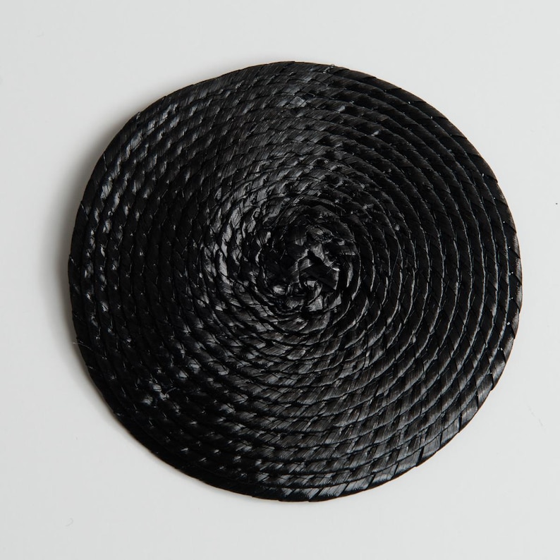 May include: A circular coaster constructed from a continuous black cord coiled in a tight spiral pattern. The surface features a textured, braided appearance created by the overlapping layers of the dark material, forming a flat, round shape.