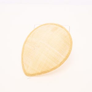 May include: A teardrop-shaped fascinator hat in a light beige, natural straw-like woven texture. The headpiece features a smooth, rounded silhouette with a finished edge, presenting a clean, structured form without any additional embellishments.