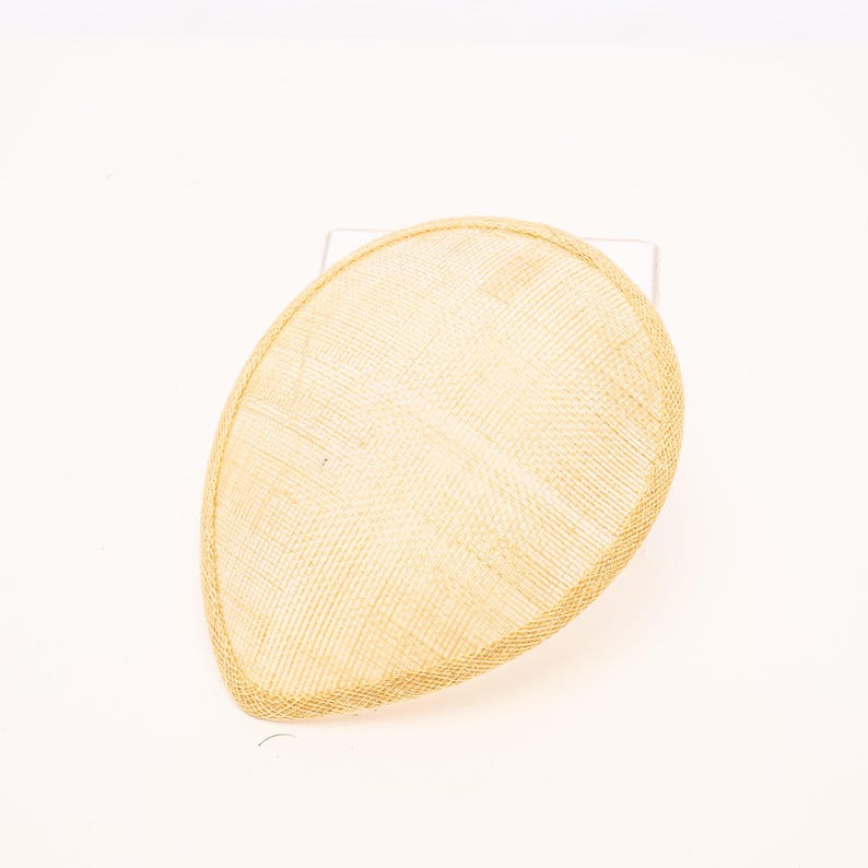 May include: A teardrop-shaped fascinator hat in a light beige, natural straw-like woven texture. The headpiece features a smooth, rounded silhouette with a finished edge, presenting a clean, structured form without any additional embellishments.