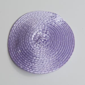 May include: A circular lavender coaster constructed from a continuous length of braided cord coiled tightly in a spiral pattern. The surface has a subtle sheen and a uniform texture created by the interlocking strands of the light purple rope.