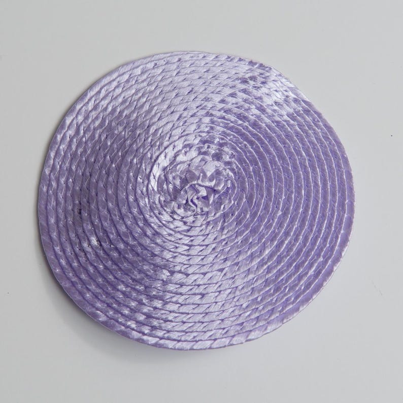 May include: A circular lavender coaster constructed from a continuous length of braided cord coiled tightly in a spiral pattern. The surface has a subtle sheen and a uniform texture created by the interlocking strands of the light purple rope.