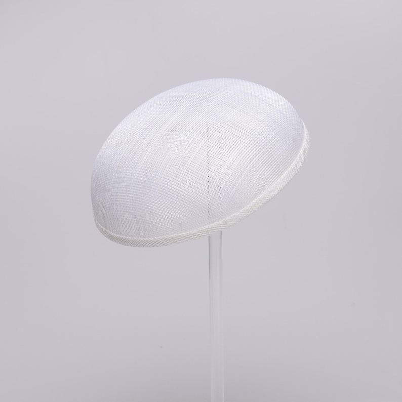 May include: A solid white, dome-shaped hat with a woven, textured surface. The hat features a clean, rounded silhouette and a finished edge, presented on a clear, vertical stand against a plain, light-colored background.