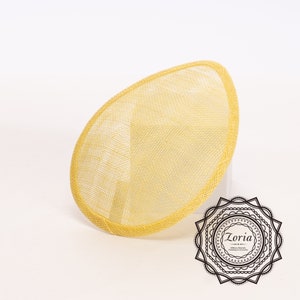 May include: A yellow oval fascinator hat base with a woven mesh texture and a finished edge. The design features a smooth, rounded silhouette. In the bottom right corner, a black circular logo displays the text Zoria Millinery, Sharrows, Trimmings & Accessories.