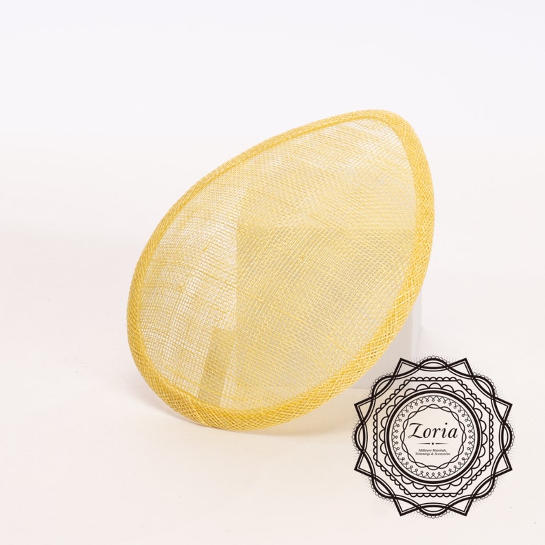 May include: A yellow oval fascinator hat base with a woven mesh texture and a finished edge. The design features a smooth, rounded silhouette. In the bottom right corner, a black circular logo displays the text Zoria Millinery, Sharrows, Trimmings & Accessories.