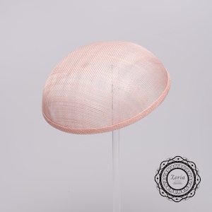 May include: A pale pink dome-shaped fascinator hat with a woven mesh texture and a finished edge. The hat is displayed on a clear stand. In the bottom right corner, there is a circular black and white logo containing the text Zoria.