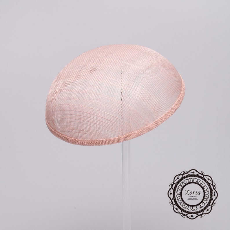 May include: A pale pink dome-shaped fascinator hat with a woven mesh texture and a finished edge. The hat is displayed on a clear stand. In the bottom right corner, there is a circular black and white logo containing the text Zoria.
