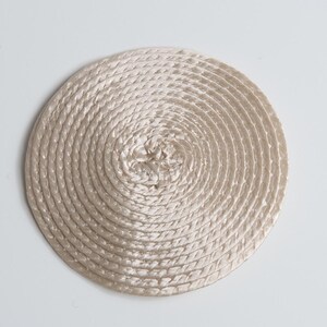 May include: A circular coaster or mat constructed from a continuous, tightly coiled beige satin cord. The material features a subtle sheen and is arranged in a spiral pattern, radiating from the center to the outer edge of the round shape.