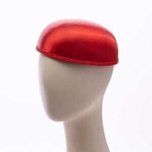 May include: A bright red, dome-shaped pillbox hat with a smooth, satin-like finish. The hat features a structured, rounded crown and a matching red braided trim along the bottom edge, providing a clean, finished look to the circular base.
