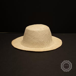 May include: A beige woven straw hat with a rounded crown and a wide brim. The hat is made of a light-colored, textured material and sits on a dark surface. The hat is a classic style, suitable for sun protection.